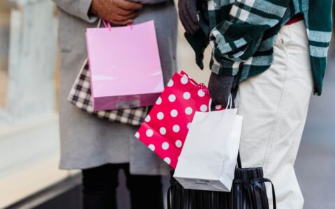 Unrecognizable female friends with various colorful shopping bags standing on sidewalk near showcase on street in city on blurred background