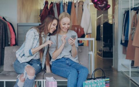 Two young women sitting in a clothing store enjoying shopping and browsing on a smartphone.