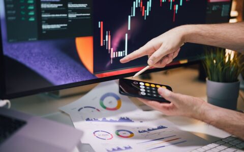 Hands analyzing stock market data on monitor and smartphone in office setting.