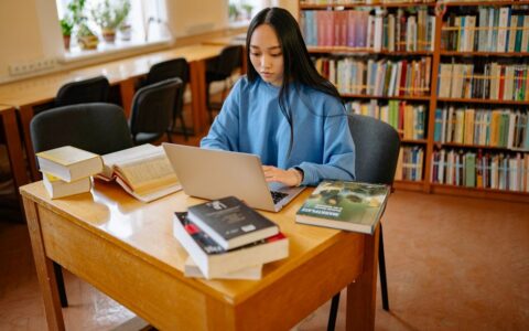 A teenager intensely focused on studying with a laptop in a library setting.