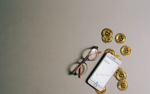 Flat lay of Bitcoin coins, smartphone with graph, and glasses on gray surface.