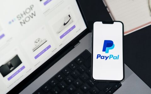 Smartphone displaying PayPal logo next to laptop with online shopping site open.